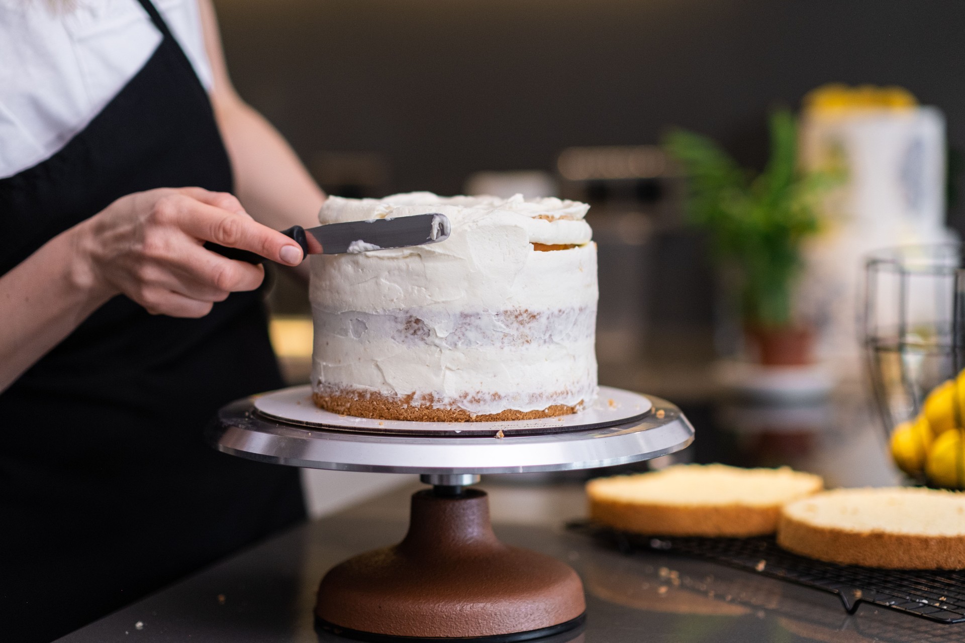 Housewife in black apron using knife to spread fresh white cream around baked layers